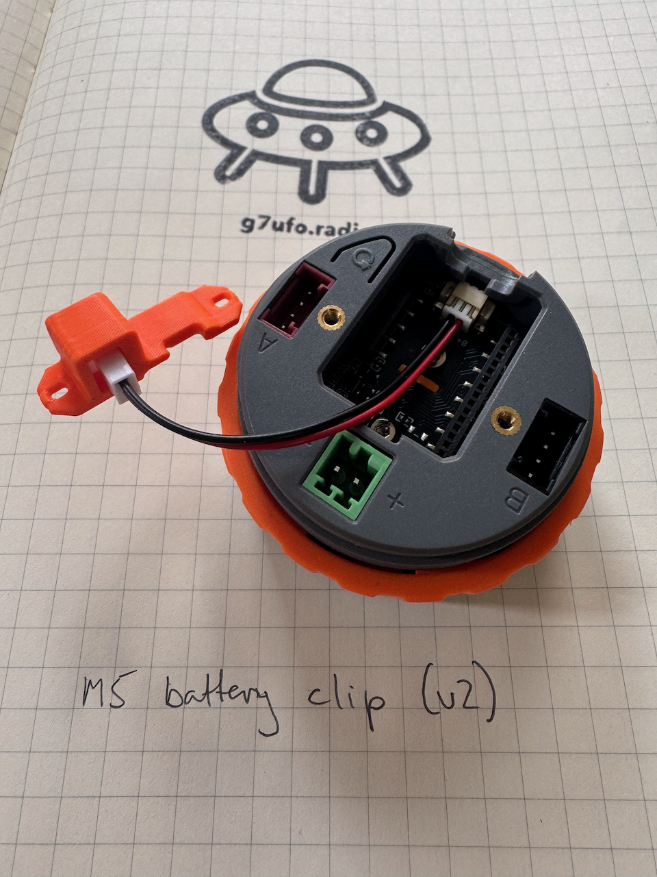 The battery clip&rsquo;s red and black cable plugged into the connector inside the M5 Dial with the STAMP S3A board removed, leading out to the orange battery adapter