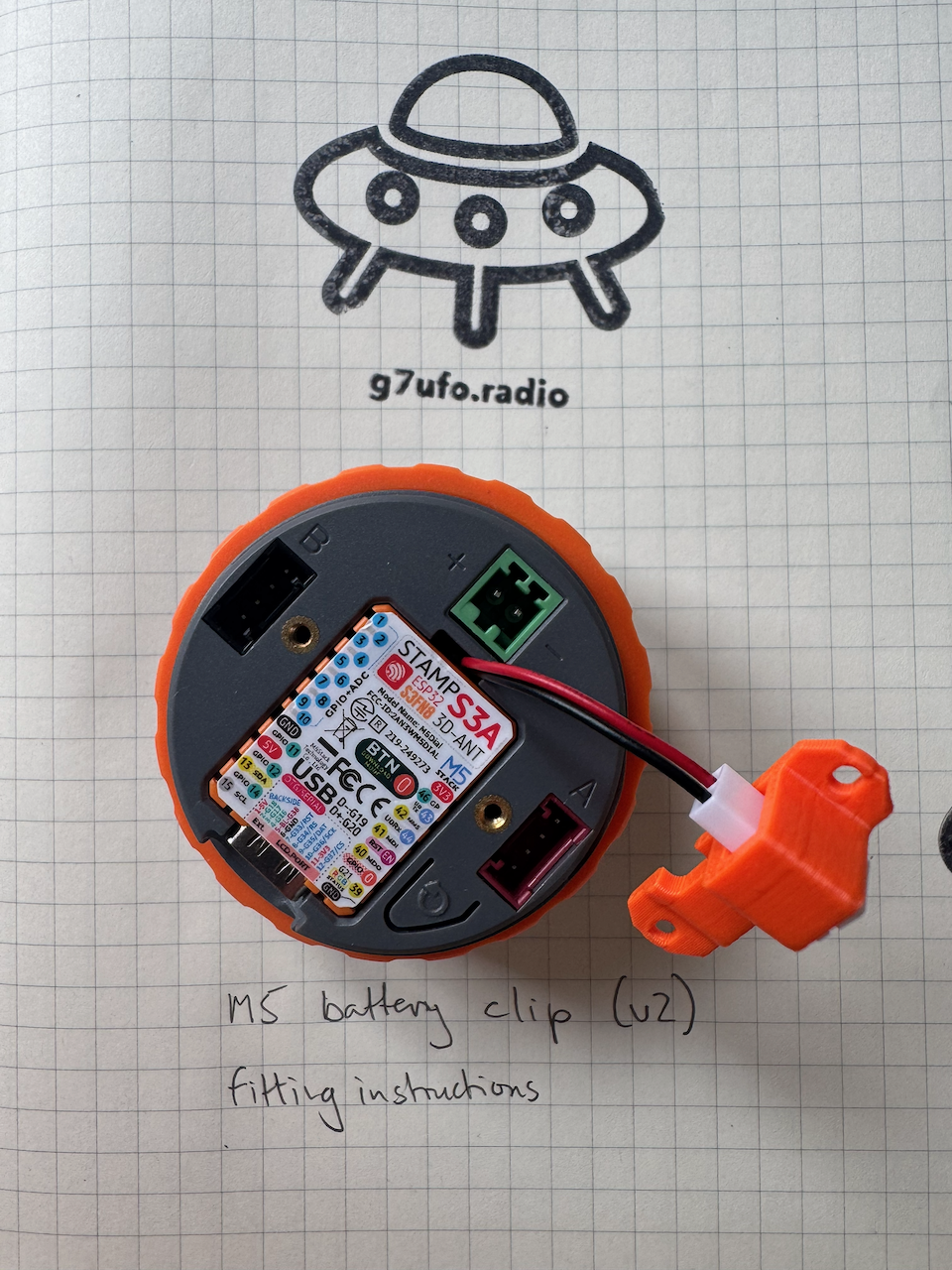 The STAMP S3A board refitted in the M5 Dial, with the battery clip&rsquo;s red and black cable running out of the groove at the top of the board to the orange battery adapter