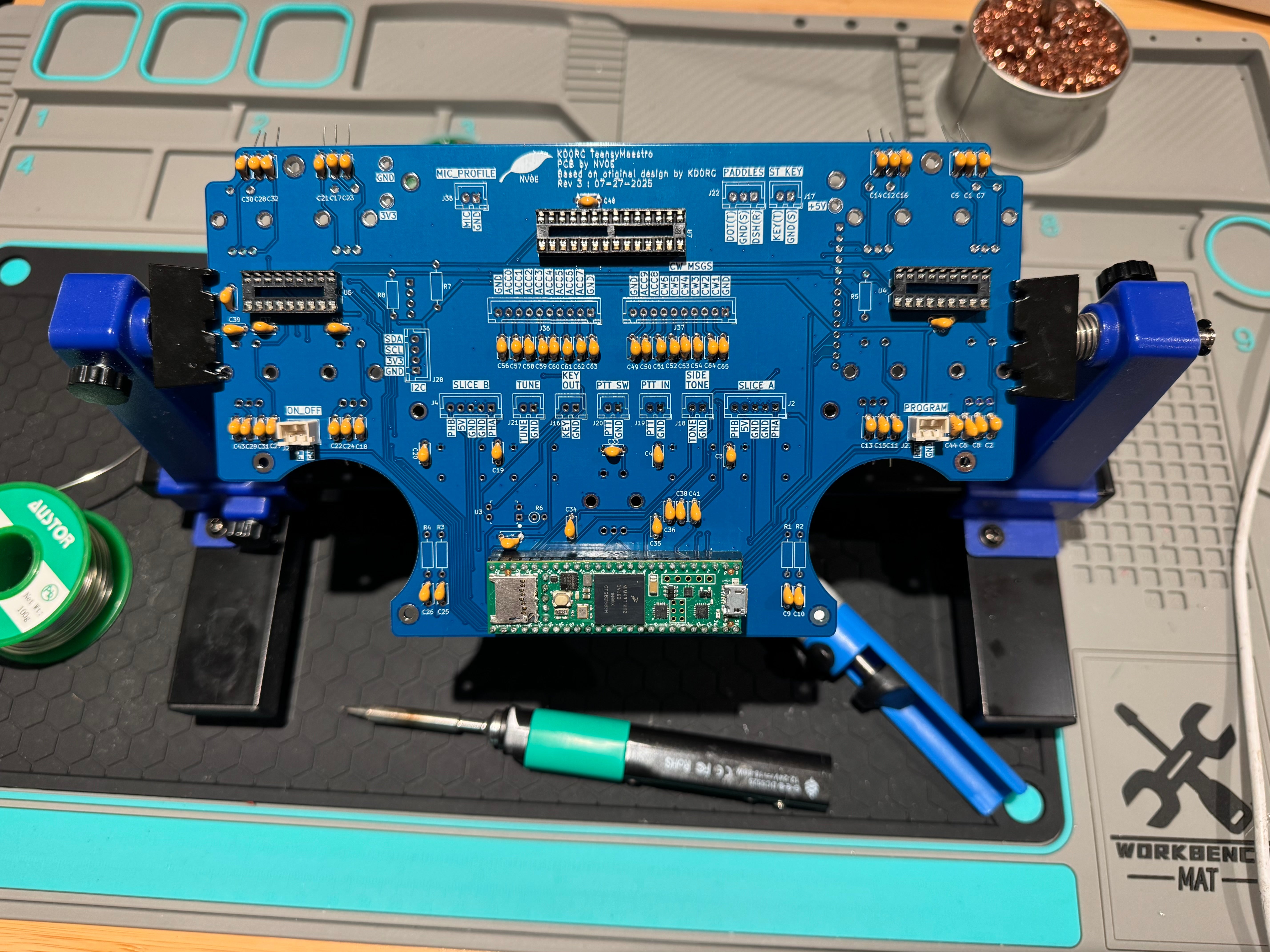 The blue TeensyMaestro PCB held in a PCB vice on a soldering mat, with the Teensy board fitted and a soldering iron alongside